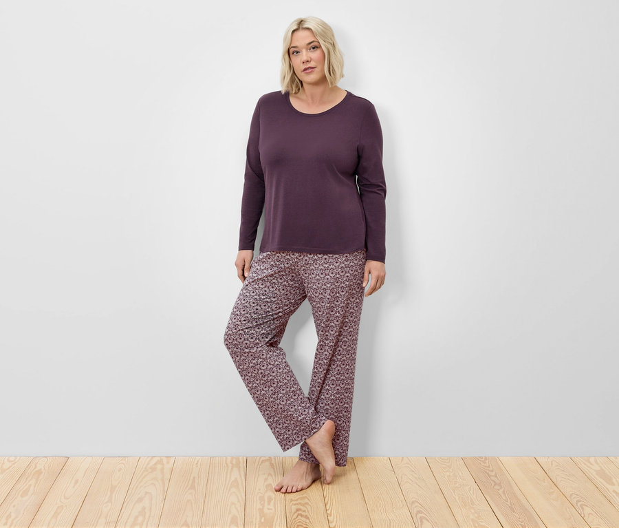 Une femme blonde en pyjama bordeaux à manches longues et pantalon à fleurs pose devant un mur sur un plancher en bois.