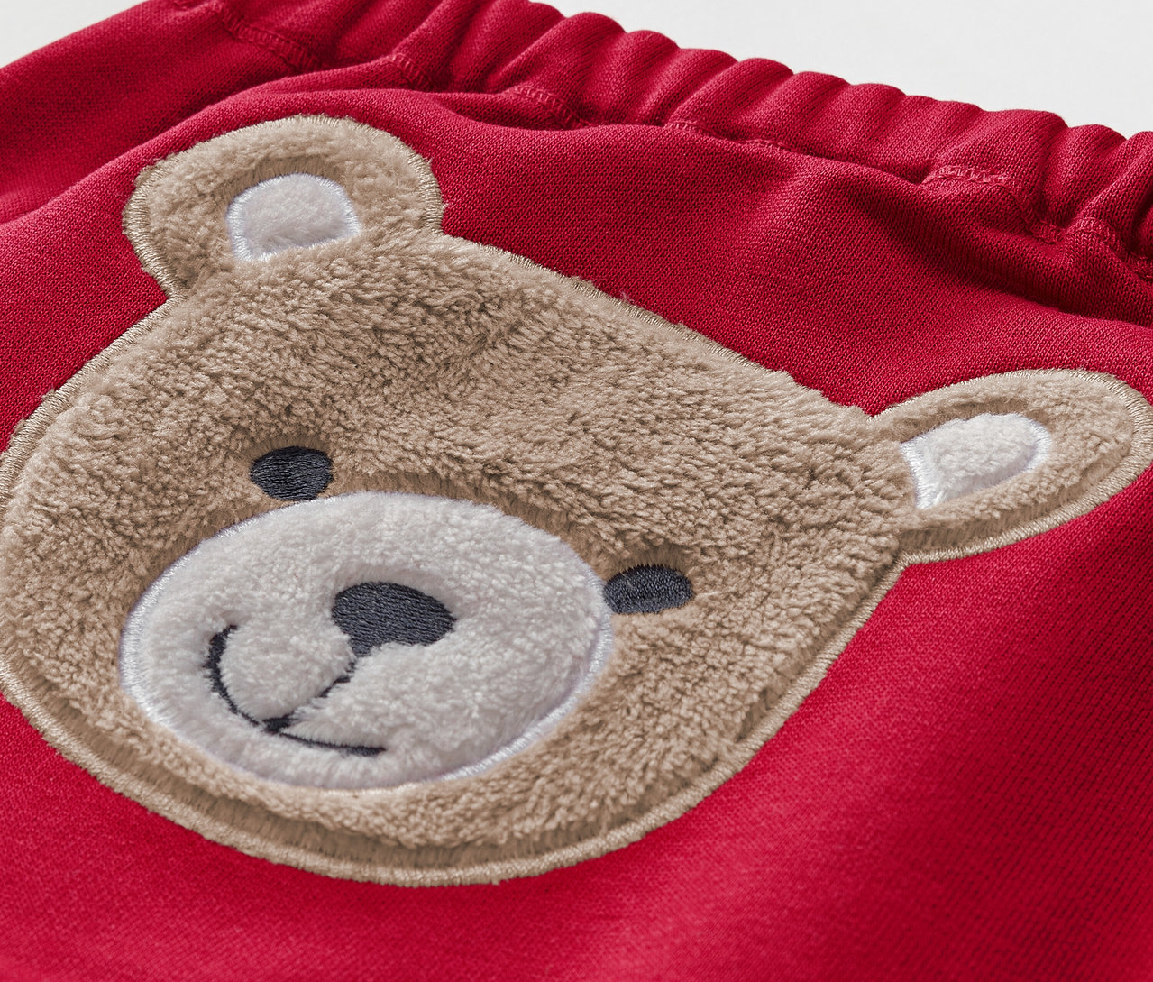 Détail d'un pantalon rouge pour enfant avec un ours en peluche marron.