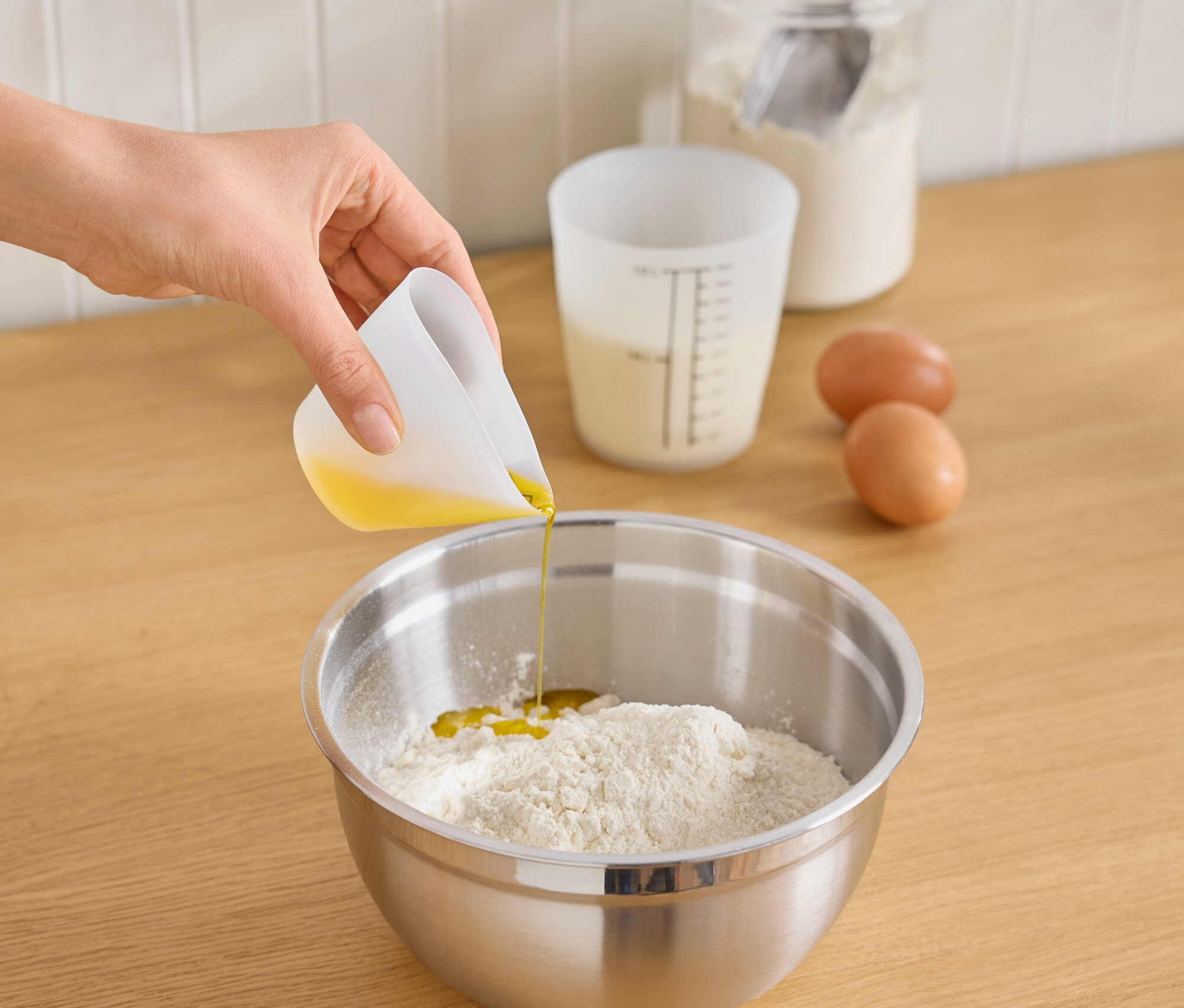 Une main verse de l'huile d'un verre doseur blanc dans un bol en acier inoxydable contenant de la farine. Sur le plan de travail se trouvent deux œufs, un verre doseur avec du lait et un pot en verre avec de la farine et une cuillère.