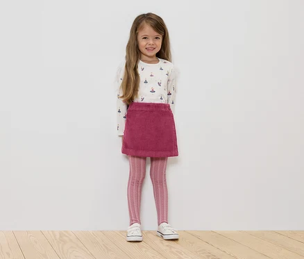Une fillette debout porte un haut blanc à manches longues avec des ballerines, une jupe en velours côtelé rose, des leggings roses à motifs et des baskets blanches.