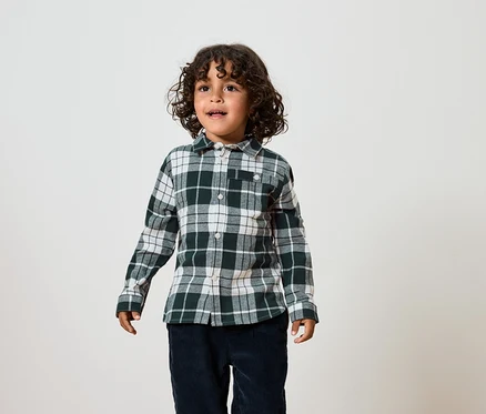 Un garçon aux cheveux bouclés porte une chemise à carreaux verte et blanche et un pantalon bleu foncé.