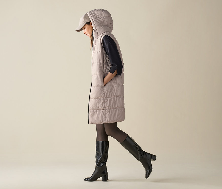 Une femme marche et porte un gilet matelassé à capuche, une casquette et des bottes cavalières.