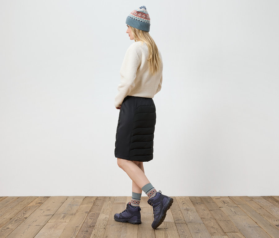 Femme posant en pull crème, jupe noire, bottes et bonnet tricoté.