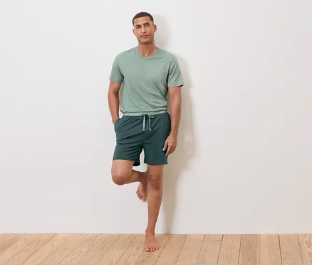Un homme se tient sur une jambe contre un mur, portant un t-shirt vert et un short.