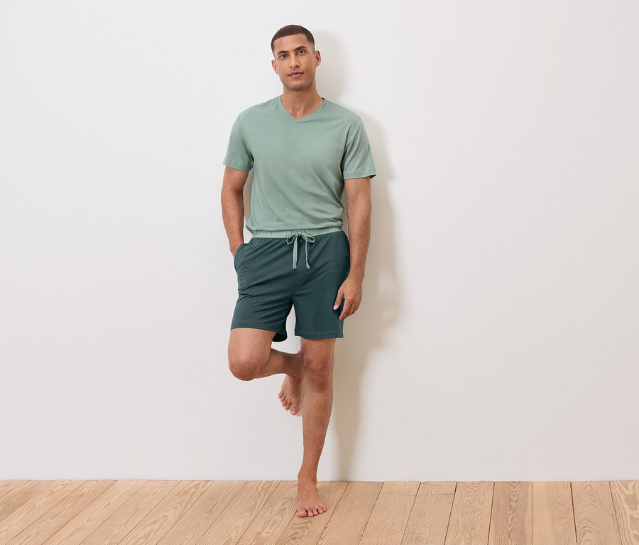 Un homme se tient sur une jambe contre un mur, portant un t-shirt vert et un short.