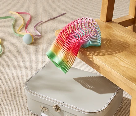 Un ressort-jouet arc-en-ciel repose partiellement sur une valise beige sous une chaise en bois.