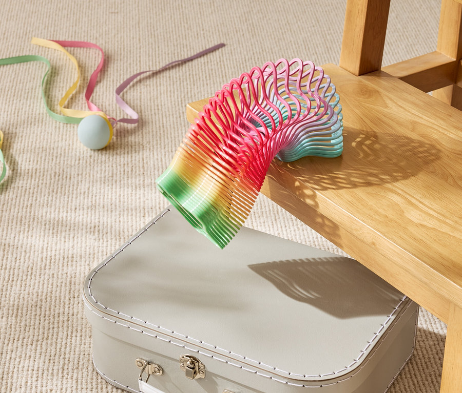 Un ressort-jouet arc-en-ciel repose partiellement sur une valise beige sous une chaise en bois.