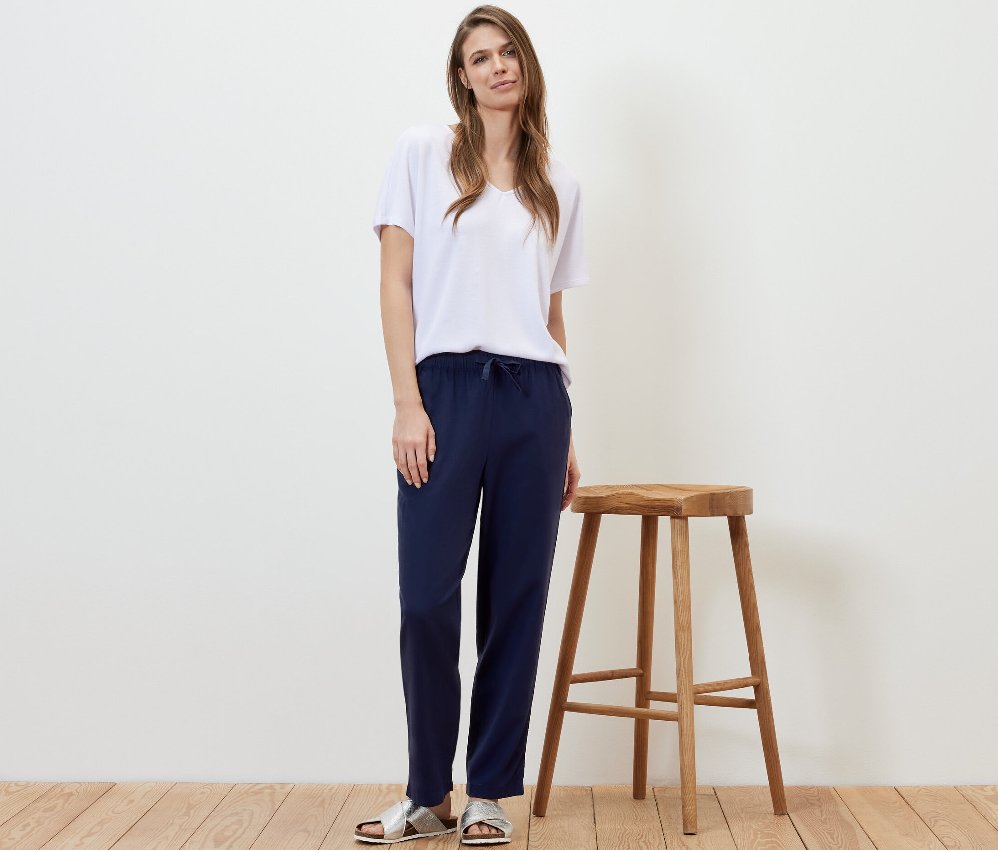 Une femme se tient à côté d'un tabouret en bois et porte un pantalon en lin bleu foncé et un t-shirt blanc.