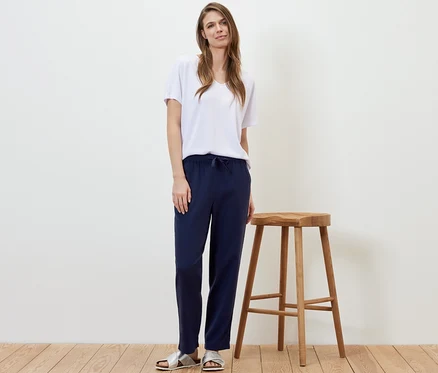Une femme se tient à côté d'un tabouret en bois et porte un pantalon en lin bleu foncé et un t-shirt blanc.