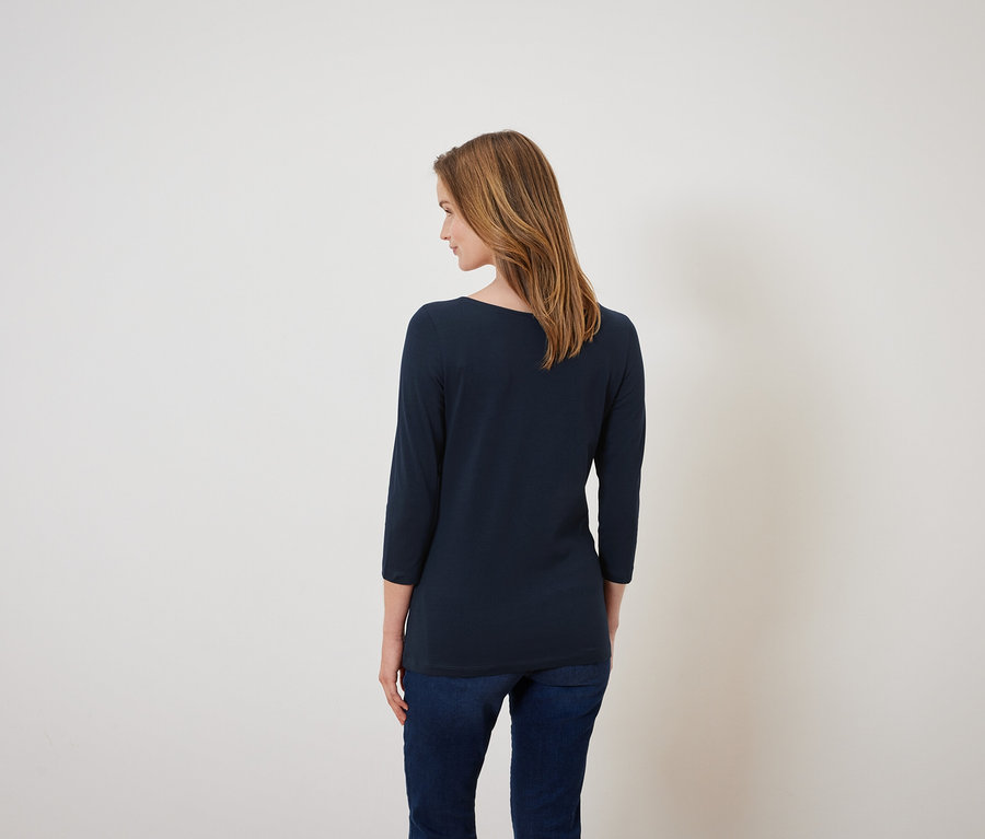 Une femme se tient dos à la caméra, portant un t-shirt bleu marine à manches trois-quarts et un jean.