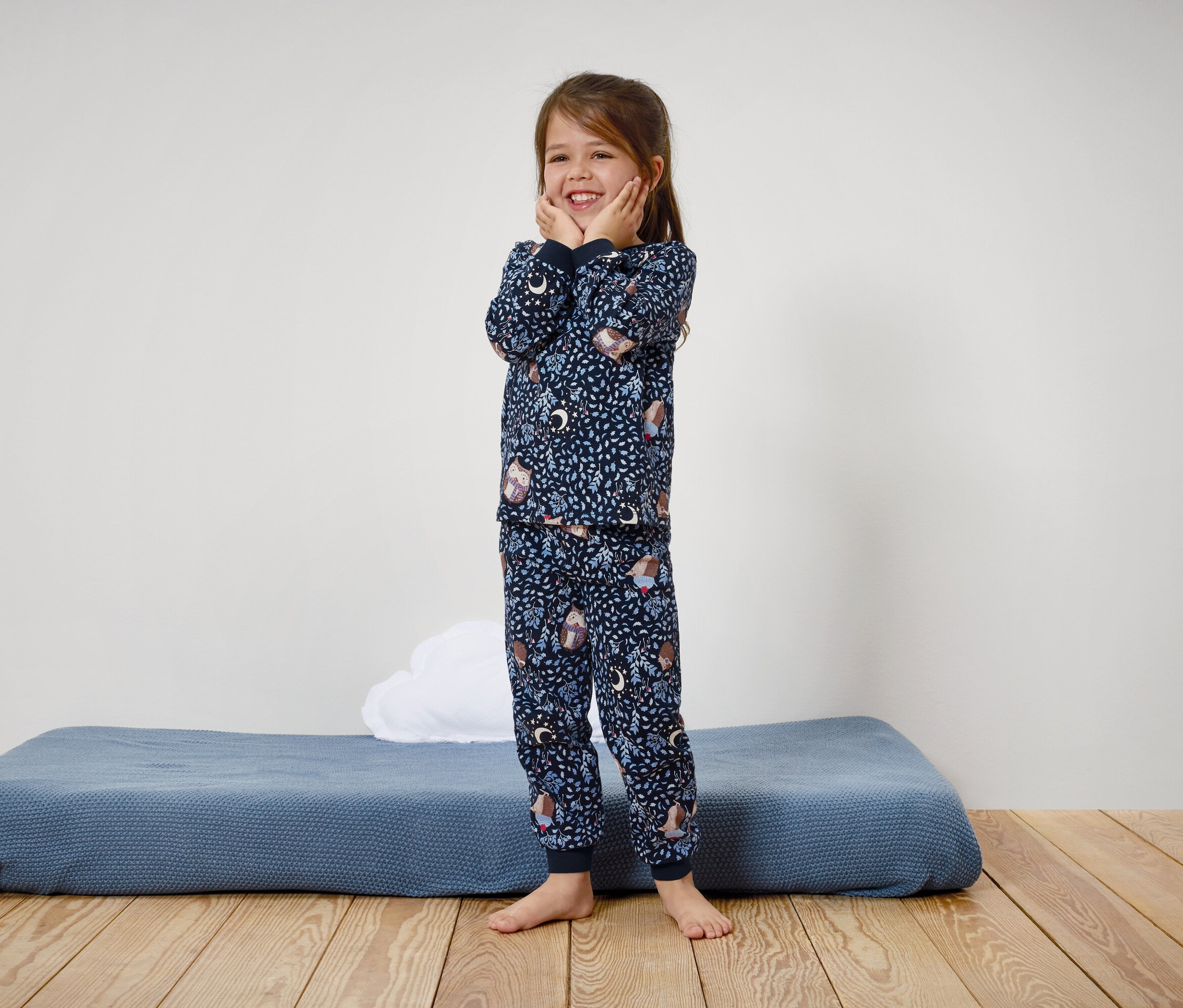 Une fillette souriante en pyjama bleu imprimé se tient sur un plancher en bois.