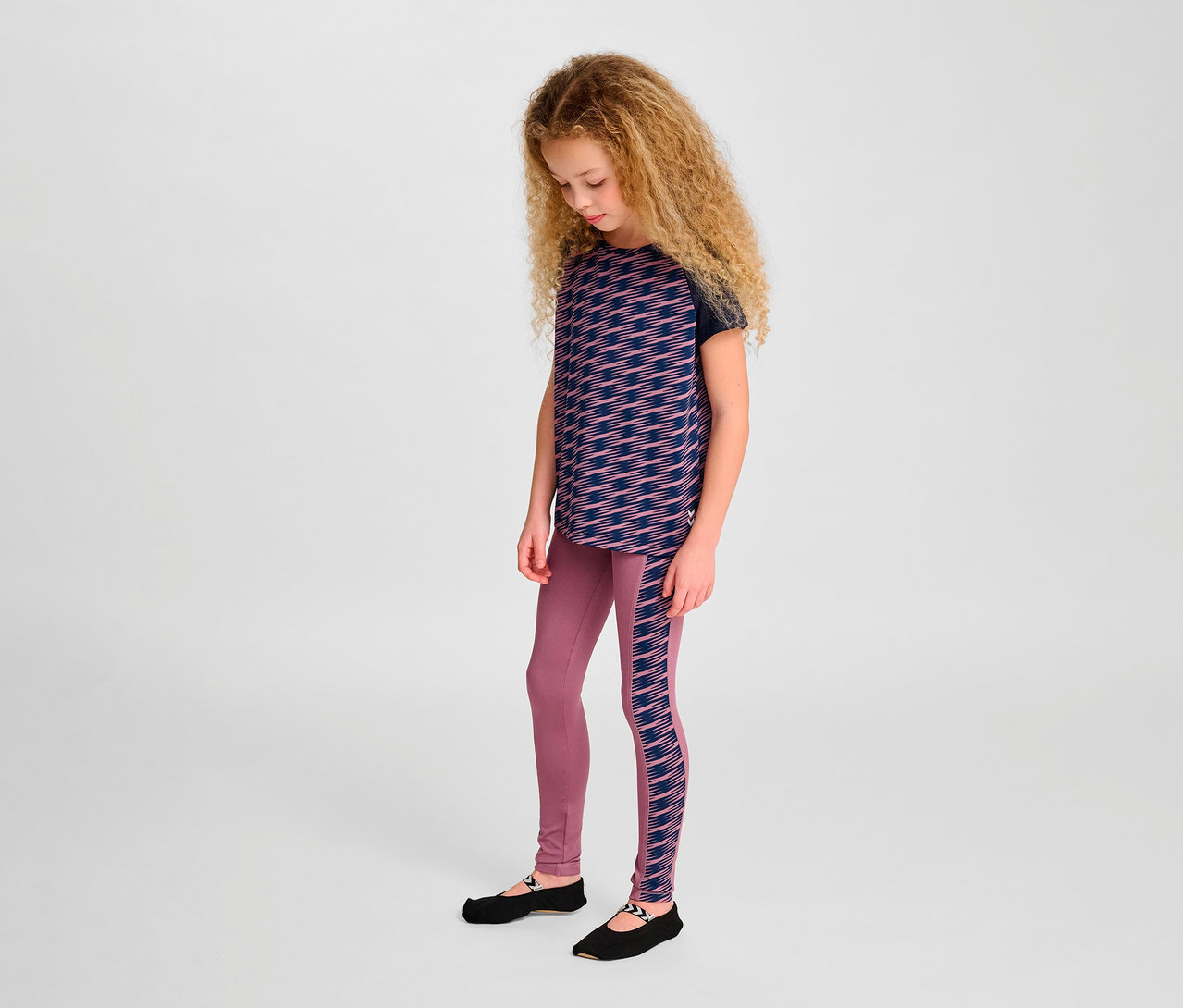 Une fille aux cheveux blonds ondulés porte un t-shirt S/S HUMMEL HMLJR Mix, un legging et des chaussures noires.