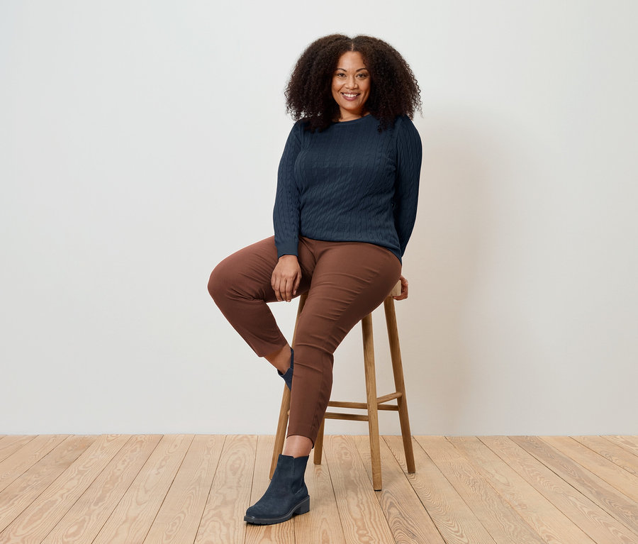 Une femme est assise sur un tabouret en bois, portant un pull bleu marine, un pantalon marron et des bottes bleues.