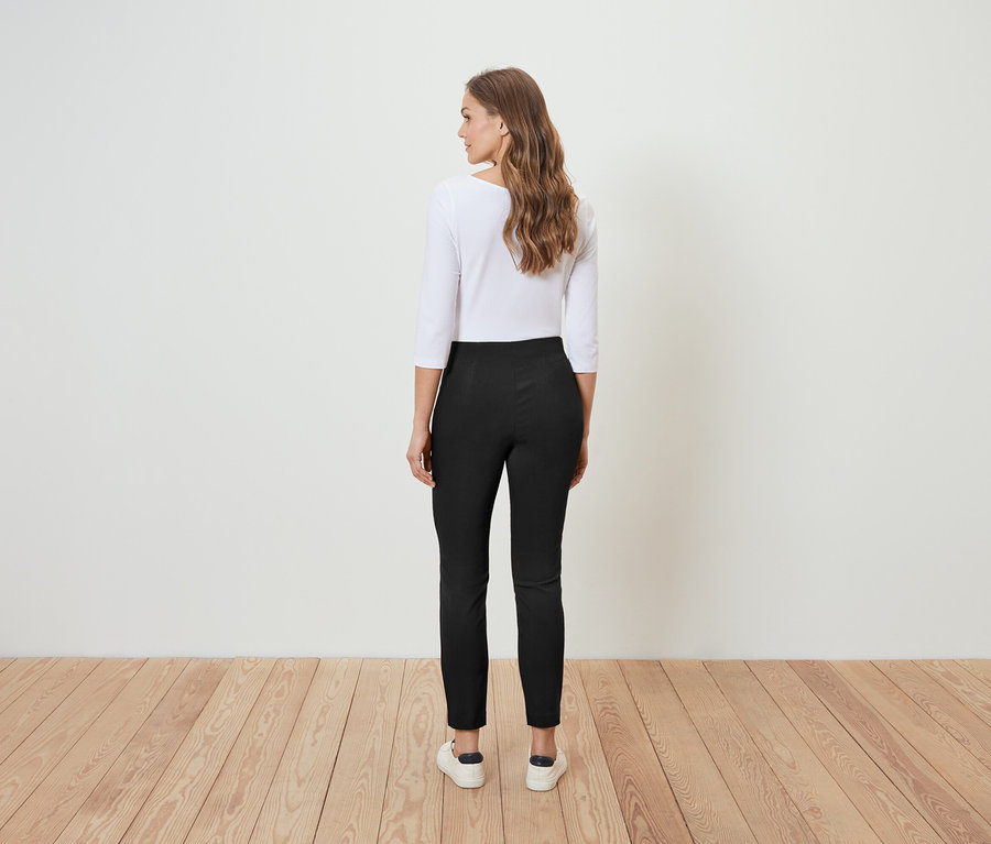 Une femme se tient dos à la caméra et porte un pantalon marron et un chemisier blanc.