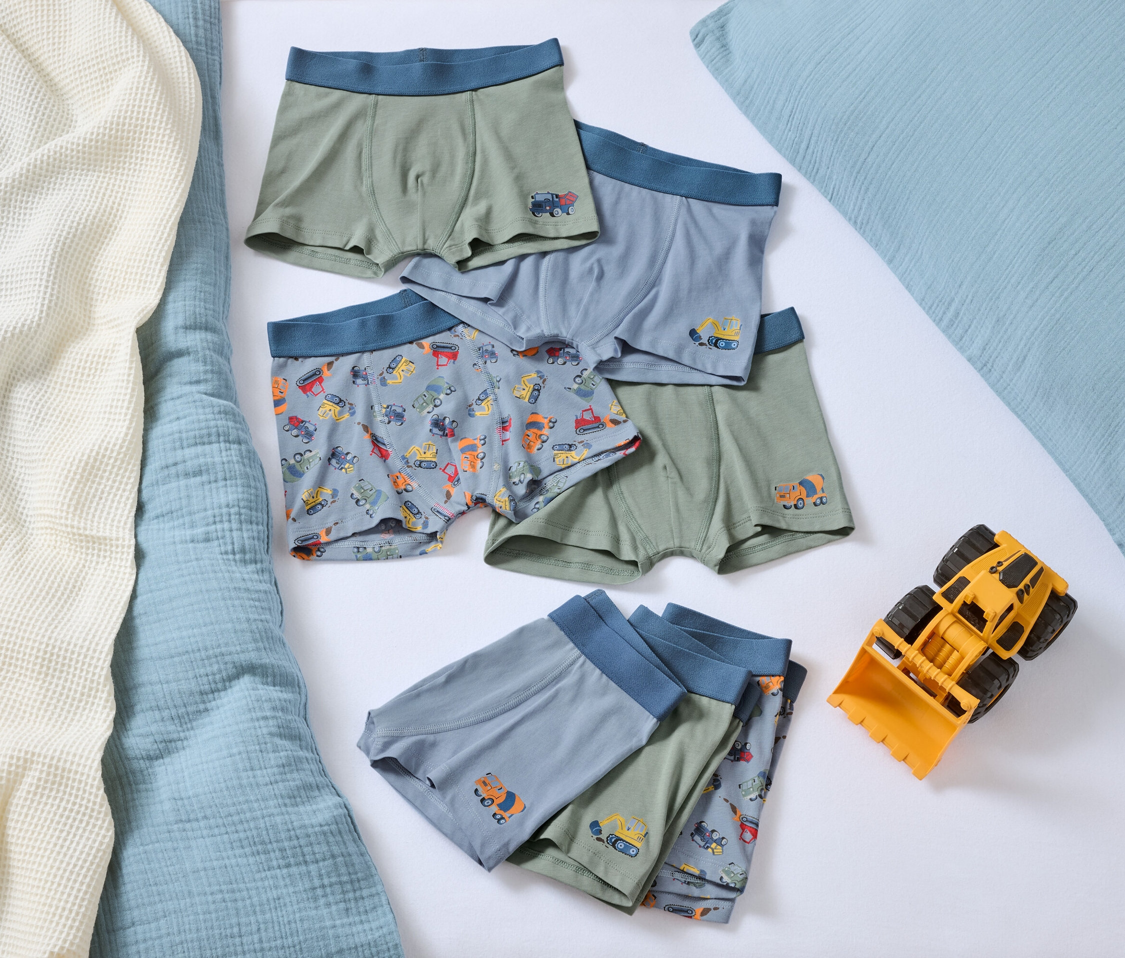 Sur un lit, plusieurs boxers pour garçons avec des motifs de voitures et un bulldozer jouet jaune.