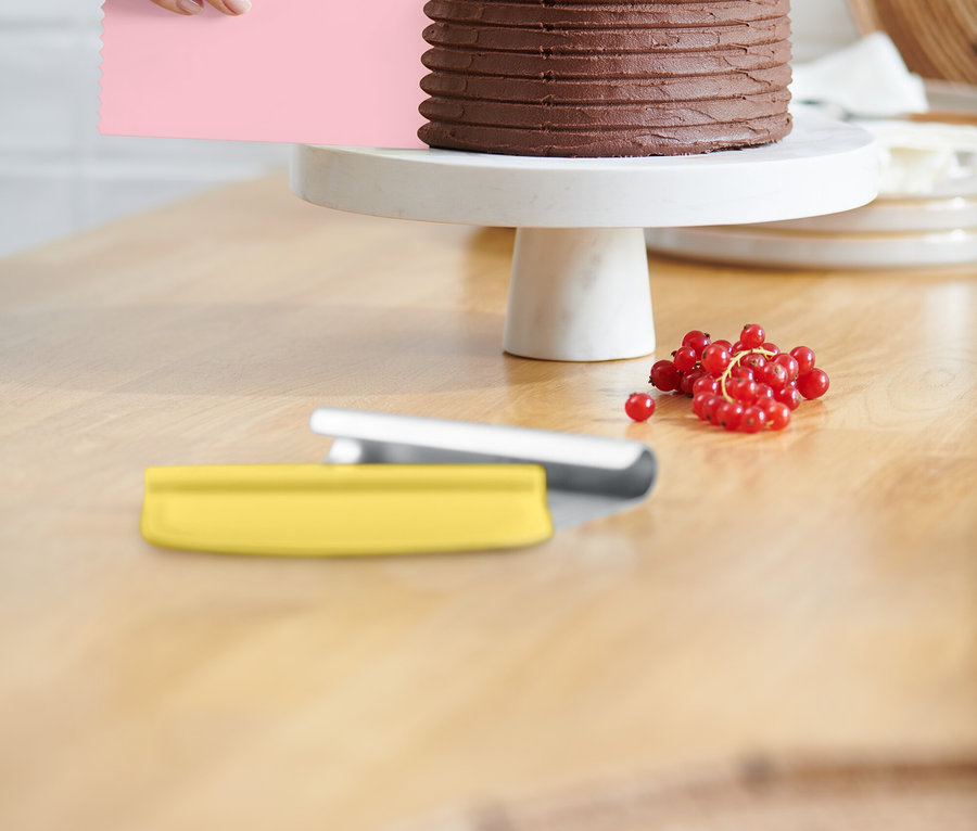 Une main lisse le glaçage d'un gâteau au chocolat avec une spatule rose. Le gâteau repose sur un présentoir blanc et est décoré de groseilles rouges. Une spatule jaune est posée sur la table.