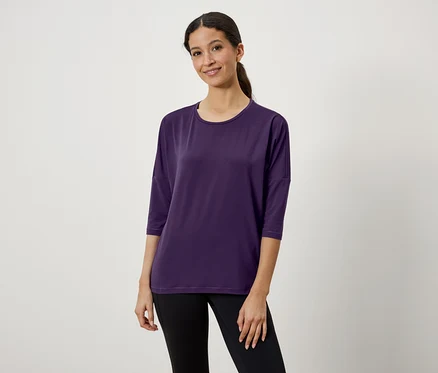 Une femme aux cheveux bruns attachés en queue de cheval porte un t-shirt violet à manches trois-quarts et un legging noir.