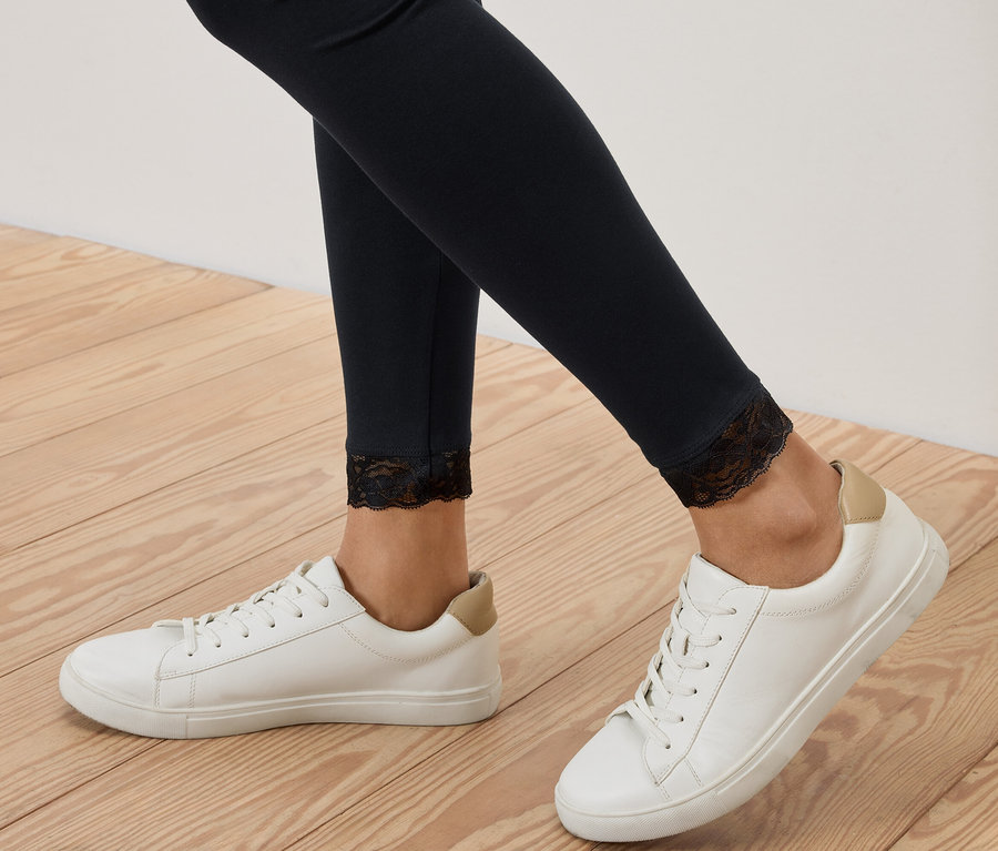 Gros plan sur des jambes portant des leggings noirs avec des finitions en dentelle, associés à des baskets blanches sur un plancher en bois.
