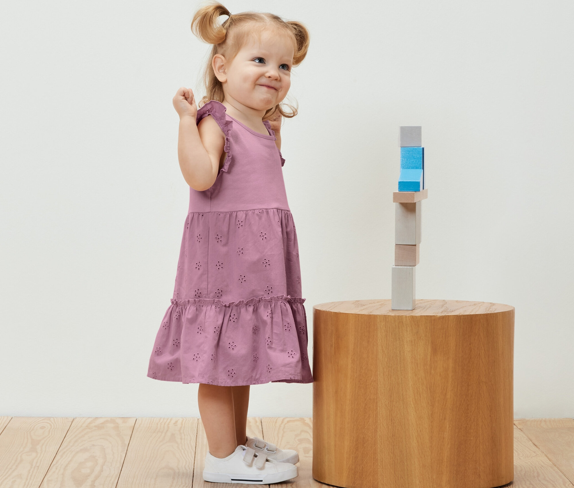 Petite fille souriante en robe pour enfant debout à côté de blocs de bois sur une table ronde.