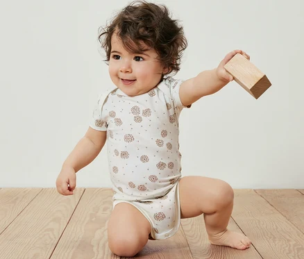 Un bébé portant un body évolutif pour bébé (lot de 5) est agenouillé sur un plancher en bois et tient un bloc de bois.