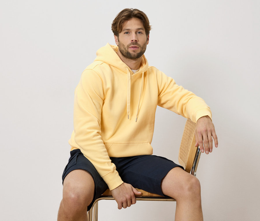 Un homme est assis sur une chaise, portant un sweat à capuche jaune et un short bleu.