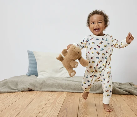 Un jeune enfant souriant aux cheveux bouclés court pieds nus sur un plancher en bois, tenant un ours en peluche et portant une combinaison blanche avec des motifs de voitures colorées.