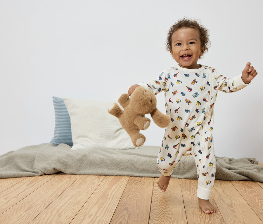 Un jeune enfant souriant aux cheveux bouclés court pieds nus sur un plancher en bois, tenant un ours en peluche et portant une combinaison blanche avec des motifs de voitures colorées.