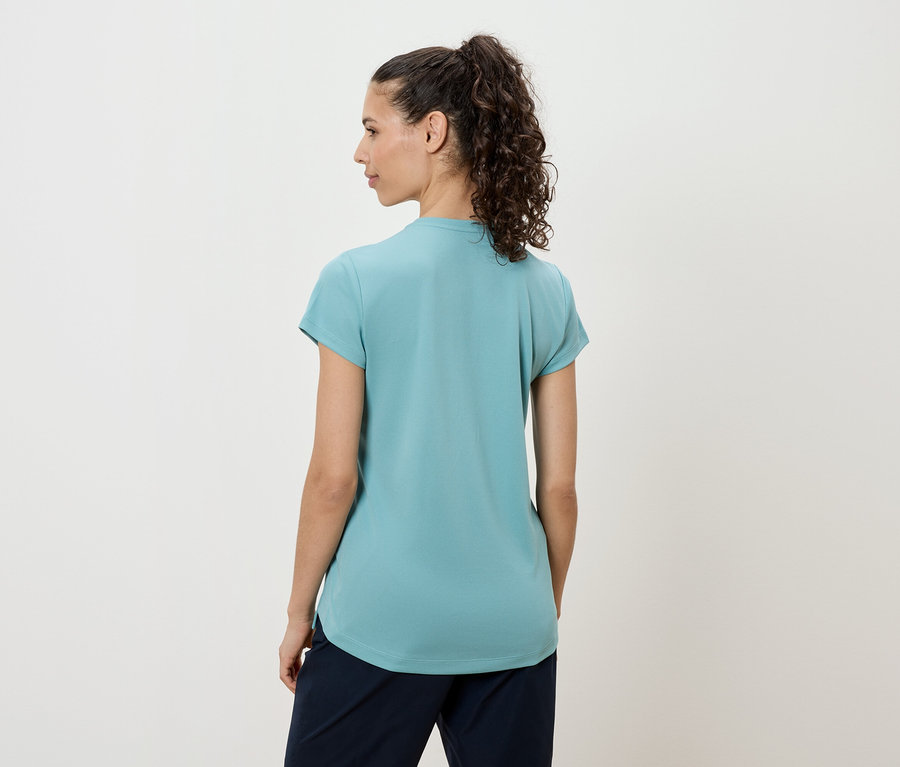 Une femme aux cheveux bouclés foncés porte un t-shirt turquoise et un pantalon foncé, vue de dos.