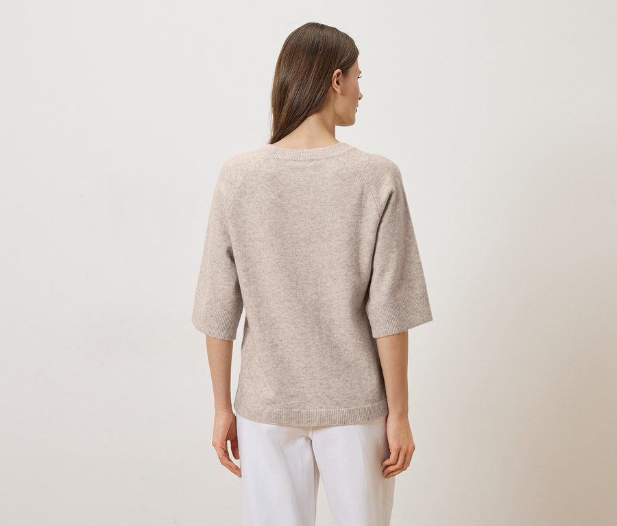 Une femme aux longs cheveux bruns porte un pull beige à manches trois quarts et un pantalon blanc, vue de dos.