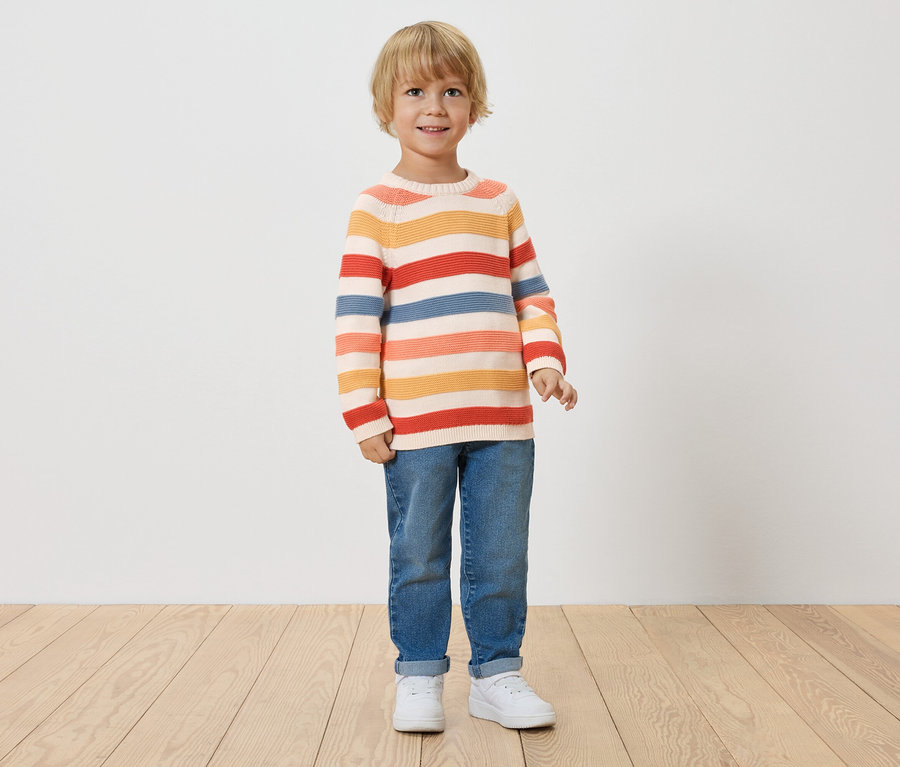 Un petit garçon aux cheveux blonds se tient devant un mur blanc, portant un pull rayé, un jean et des baskets blanches.