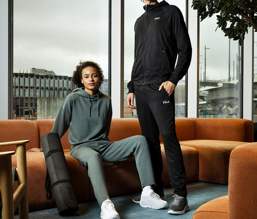 Une femme assise sur un canapé avec un hoodie pour femme FILA en mélange de modal et un tapis de yoga. Un homme se tient à côté en survêtement.