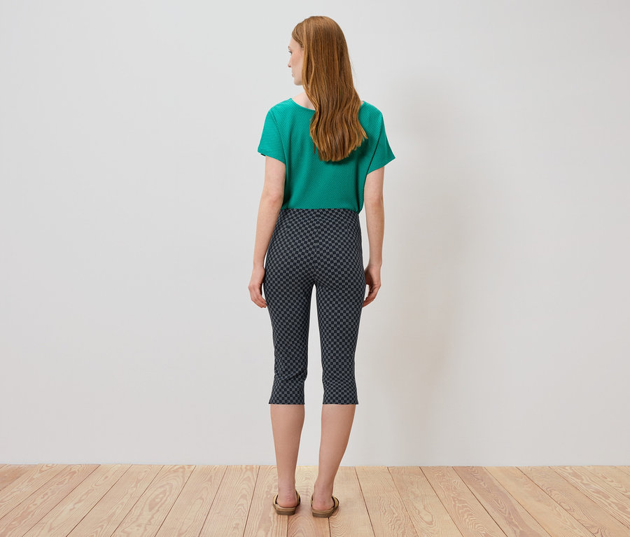 Vue arrière d'une femme aux longs cheveux roux portant un t-shirt vert, un pantacourt à motifs et des sandales.