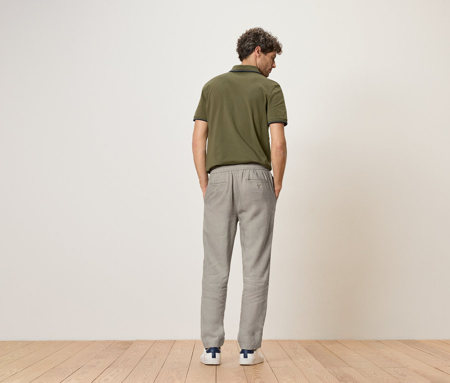 Un homme aux cheveux bouclés se tient dos à la caméra, portant un polo vert olive, un pantalon en lin gris et des chaussures blanches avec des détails bleus.