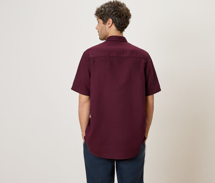 Un homme se tient dos à la caméra, portant une chemise bordeaux à manches courtes et un pantalon bleu.