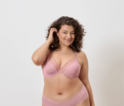 Une femme aux cheveux bouclés porte un soutien-gorge rose avec des détails en dentelle et des sous-vêtements assortis sur un fond neutre.
