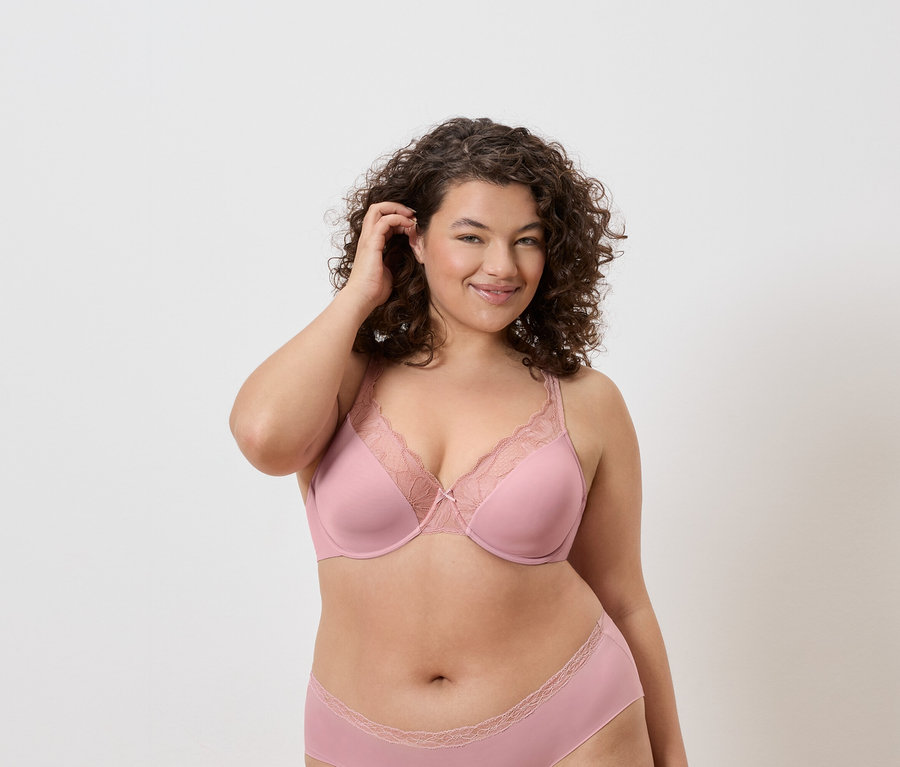 Une femme aux cheveux bouclés porte un soutien-gorge rose avec des détails en dentelle et des sous-vêtements assortis sur un fond neutre.