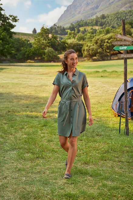 Une femme marche dans l'herbe dans une robe chemise verte avec une ceinture. On aperçoit une tente et un panneau indicateur en arrière-plan.