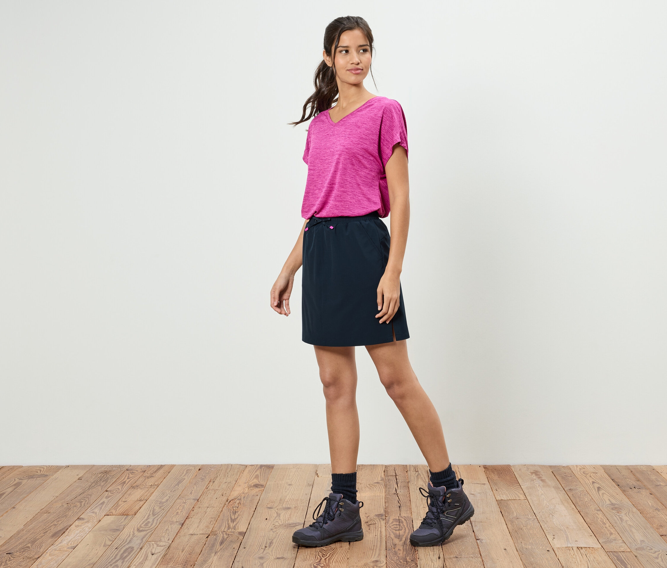 Une femme se tient devant un mur blanc portant un t-shirt rose, une jupe noire et des chaussures de randonnée.