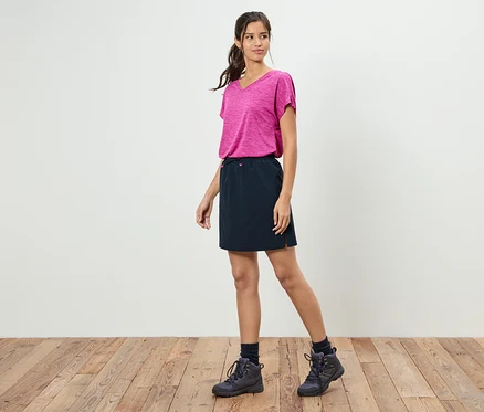 Une femme se tient devant un mur blanc portant un t-shirt rose, une jupe noire et des chaussures de randonnée.