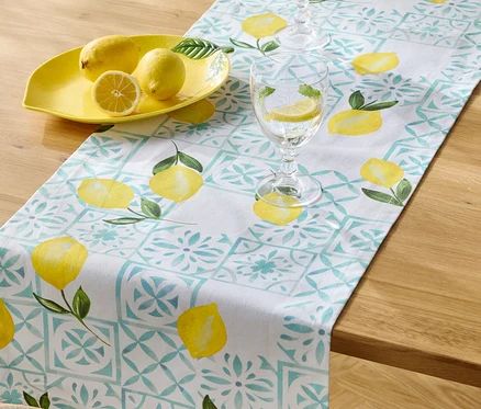 Sur une table en bois se trouvent un chemin de table à motif de citron, un bol à citron jaune avec trois citrons et deux verres d'eau avec des tranches de citron.