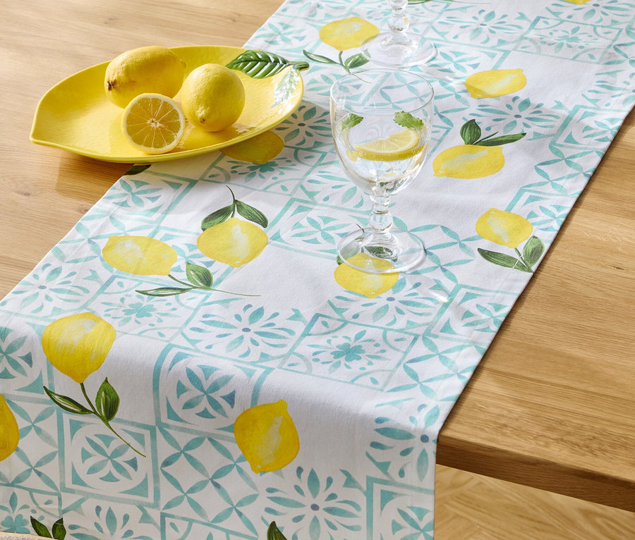 Sur une table en bois se trouvent un chemin de table à motif de citron, un bol à citron jaune avec trois citrons et deux verres d'eau avec des tranches de citron.