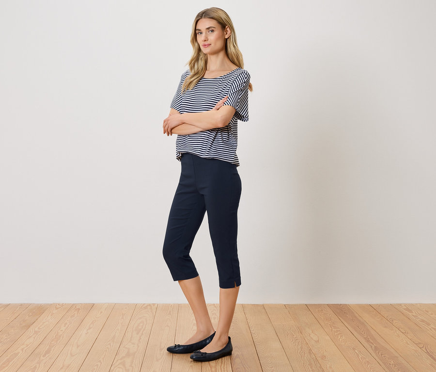 Femme aux bras croisés, portant un haut rayé bleu et blanc, un pantacourt bleu et des ballerines.