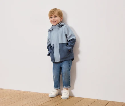 Un garçon blond souriant se tient devant un mur blanc, portant une veste bicolore bleu clair et bleu marine, un jean bleu et des baskets blanches.
