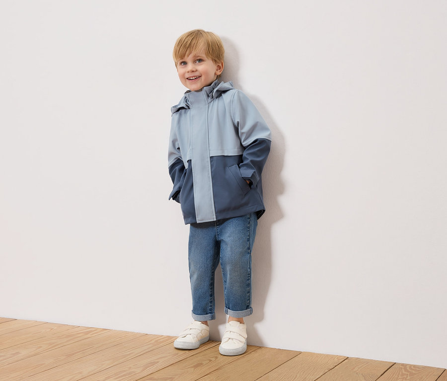 Un garçon blond souriant se tient devant un mur blanc, portant une veste bicolore bleu clair et bleu marine, un jean bleu et des baskets blanches.