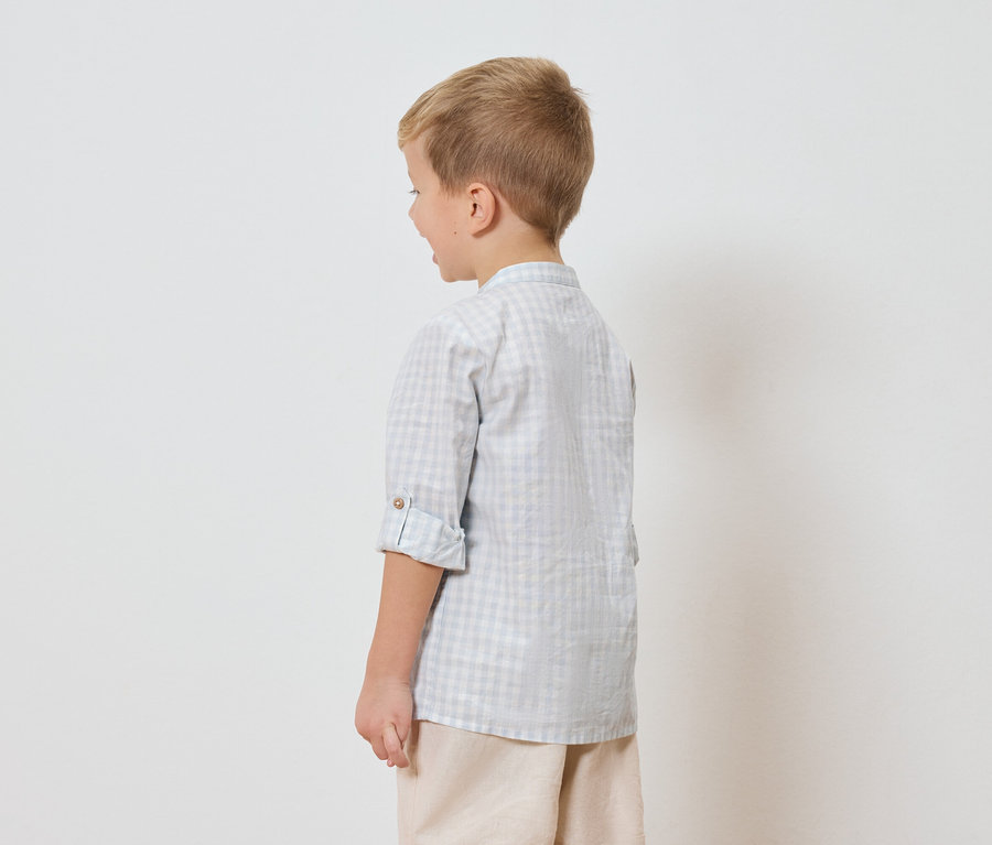 Un garçon aux cheveux blonds, vu de dos, porte une chemise à carreaux et un short beige sur fond blanc.