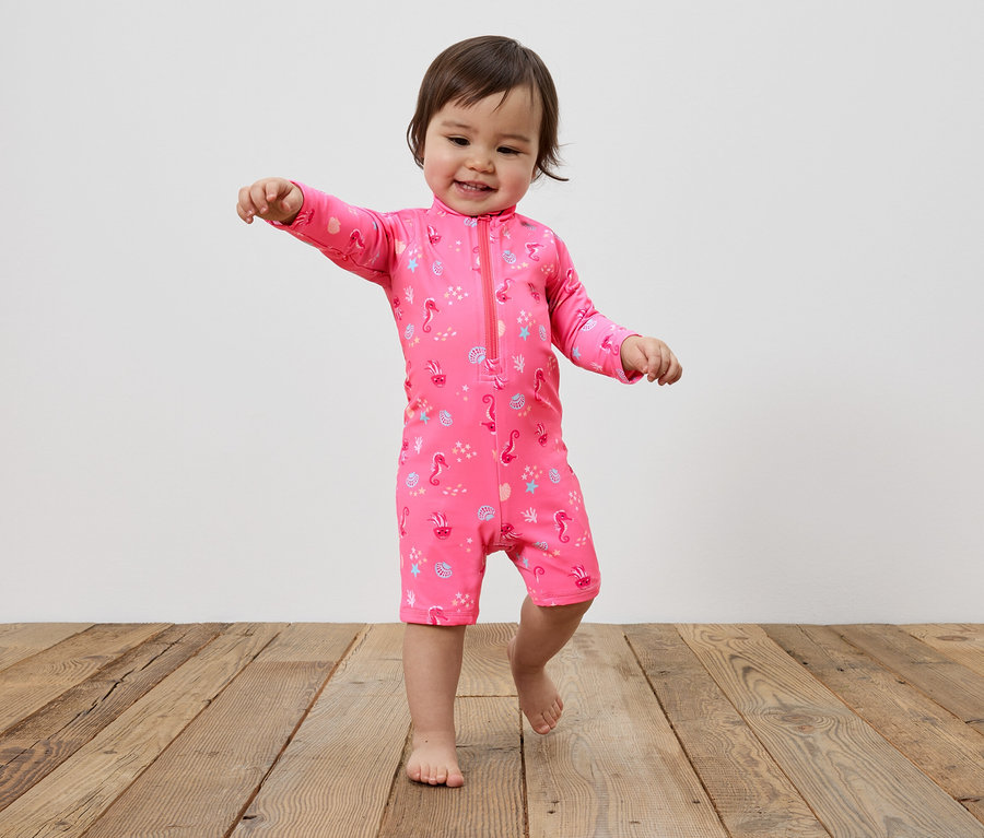 Un jeune enfant marche pieds nus sur un plancher en bois, vêtu d'un maillot de bain rose avec une fermeture éclair et un motif d'hippocampes, de coquillages et d'étoiles de mer.