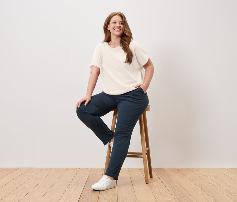 Femme assise sur un tabouret, portant un t-shirt crème, un pantalon bleu et des baskets blanches.