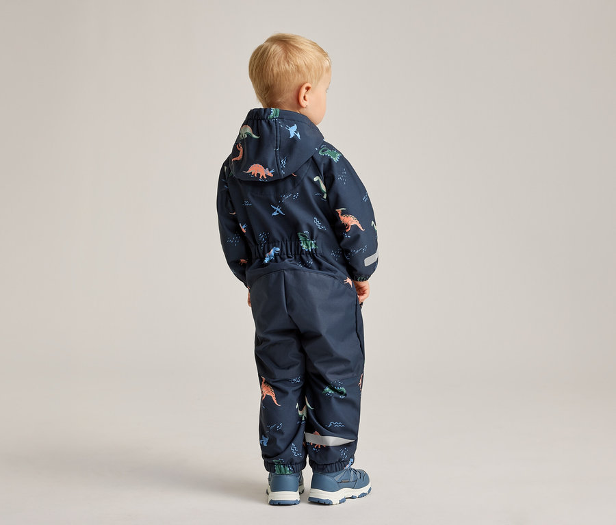 Un enfant, vu de dos, porte une combinaison bleu marine à motifs de dinosaures et des chaussures bleues.