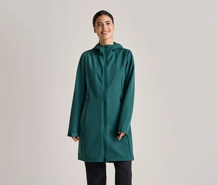Une femme souriante portant un manteau long softshell vert émeraude à capuche et un pantalon noir sur un fond gris clair.