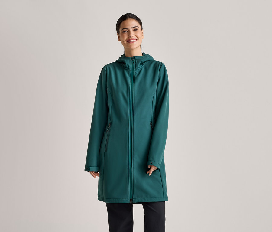 Une femme souriante portant un manteau long softshell vert émeraude à capuche et un pantalon noir sur un fond gris clair.